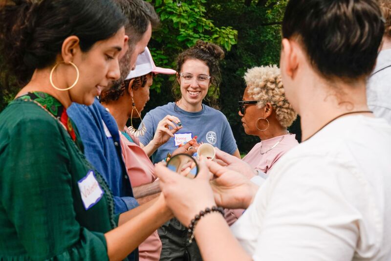 A group of people are gathered outdoors, seemingly engaged in a learning activity. A woman in a green top is holding a magnifying glass, while others around her are looking at something in her hands. The setting appears to be a park or garden, with lush greenery in the background. The atmosphere seems friendly and educational.